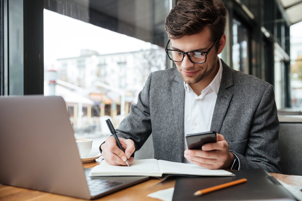 Controle de estoque - Homem sentado em mesa de escritório escrevendo anotações enquanto segura o celular